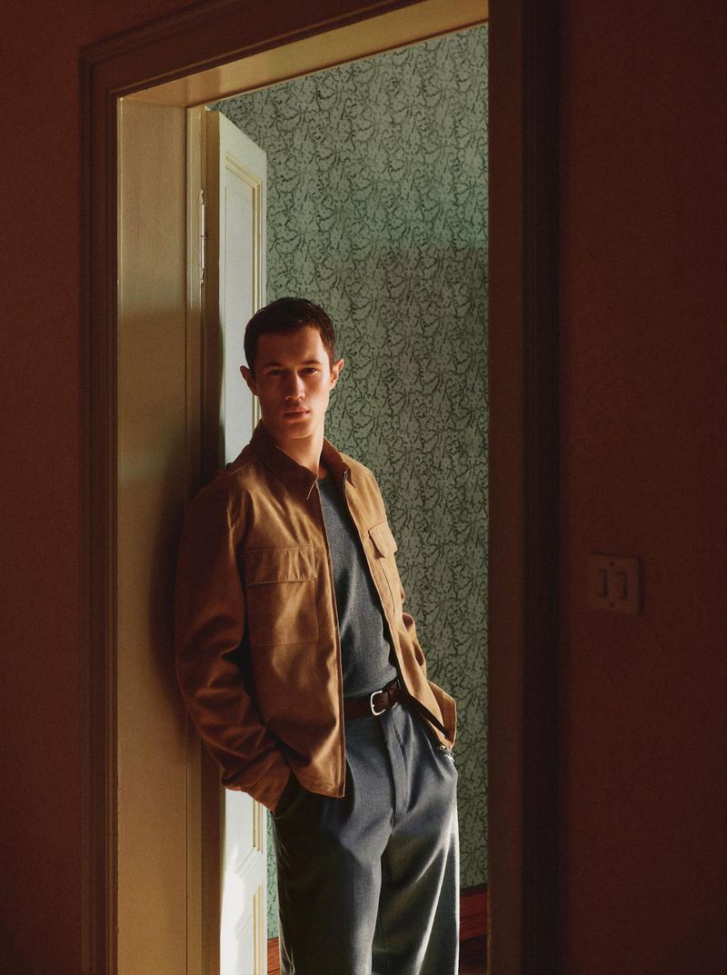 Portrait of a young adult man in a brown jacket leaning against a doorway with patterned wallpaper in the background, looking at camera.