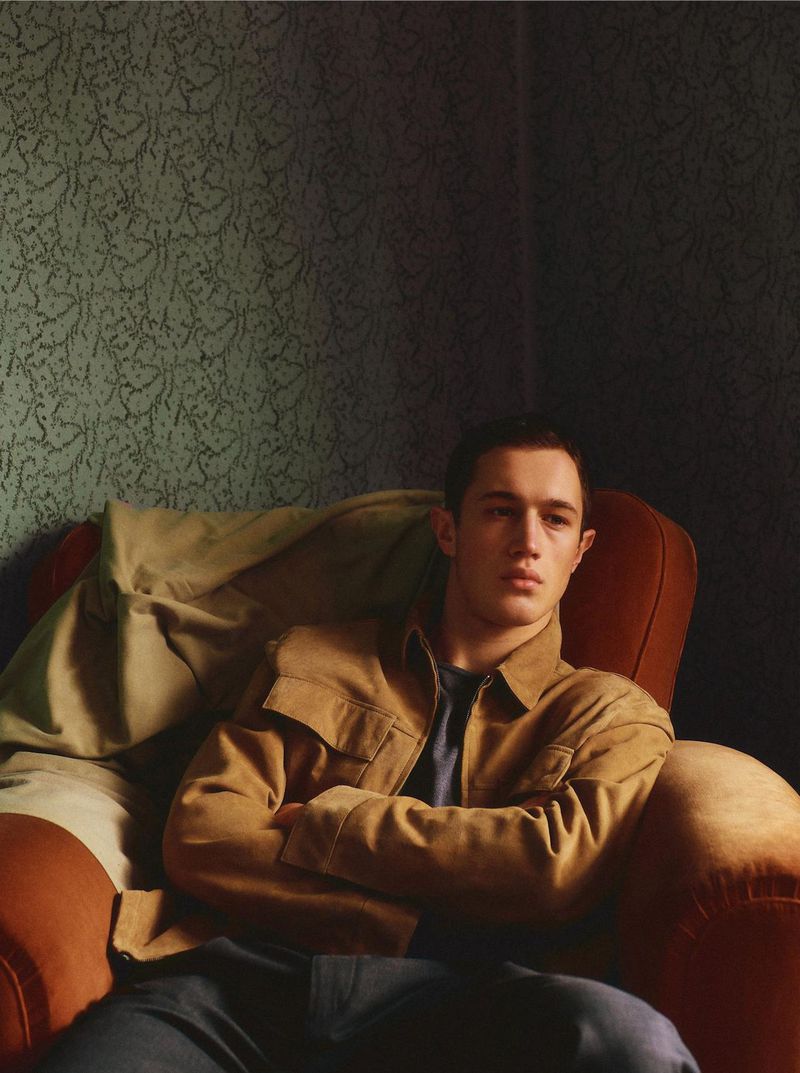 A young adult male sits in a red chair, arms crossed, wearing a tan jacket and dark pants against a muted green patterned wall.
