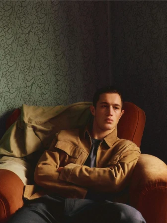 A young adult male sits in a red chair, arms crossed, wearing a tan jacket and dark pants against a muted green patterned wall.