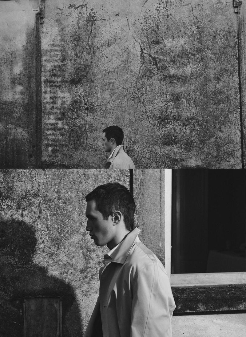 Monochrome profile of a young adult male in a beige trench coat, standing outdoors against a weathered, textured wall and window frame.