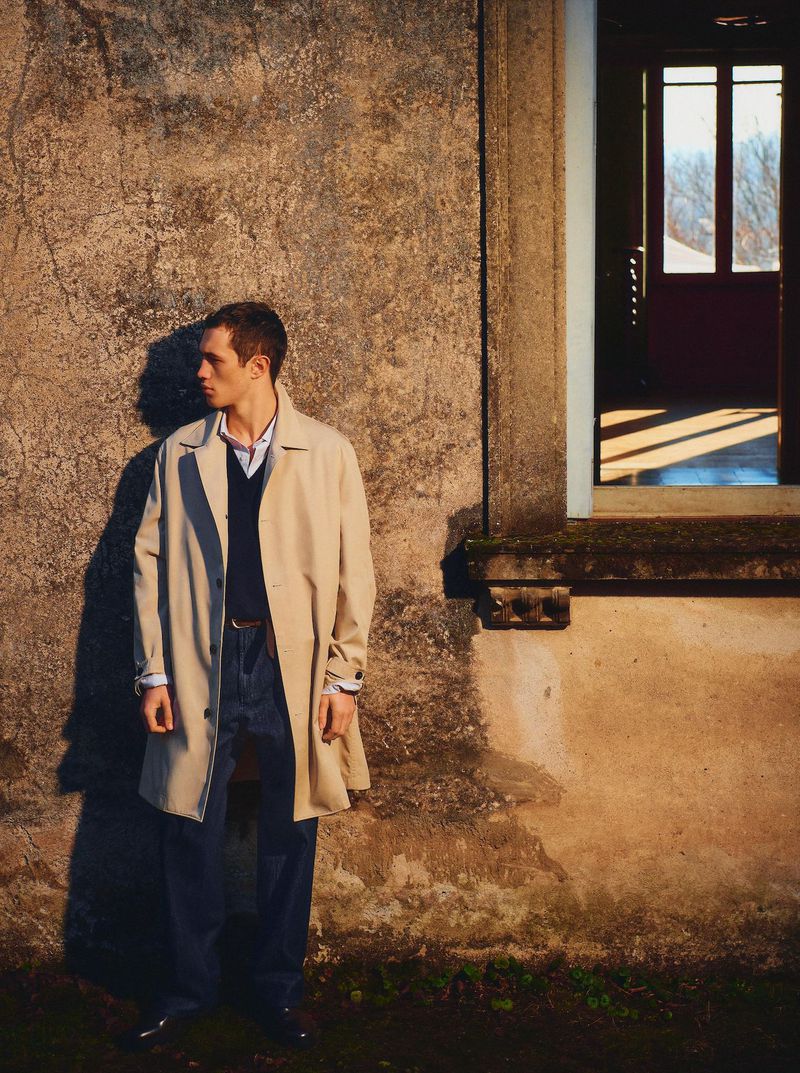 Portrait of a young man in a trench coat, standing outside a weathered building by a window, with natural sunlight casting shadows.