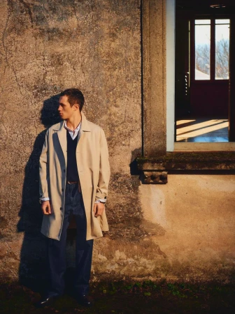 Portrait of a young man in a trench coat, standing outside a weathered building by a window, with natural sunlight casting shadows.