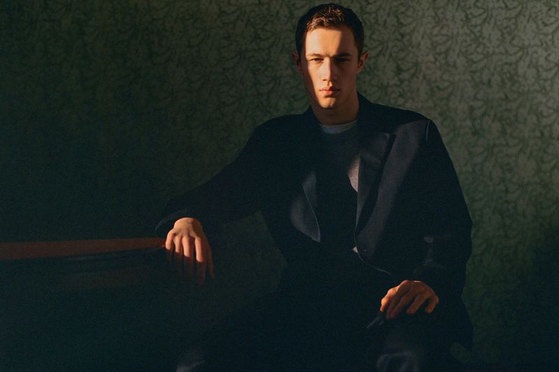 Portrait of a young adult man in a dark suit, bathed in soft light against a patterned wallpaper backdrop, conveying a sense of quiet confidence.