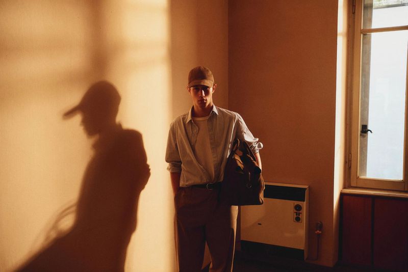 Indoor portrait of a young adult male wearing a cap, light-blue button-down shirt, and brown pants, carrying a bag, with soft lighting.
