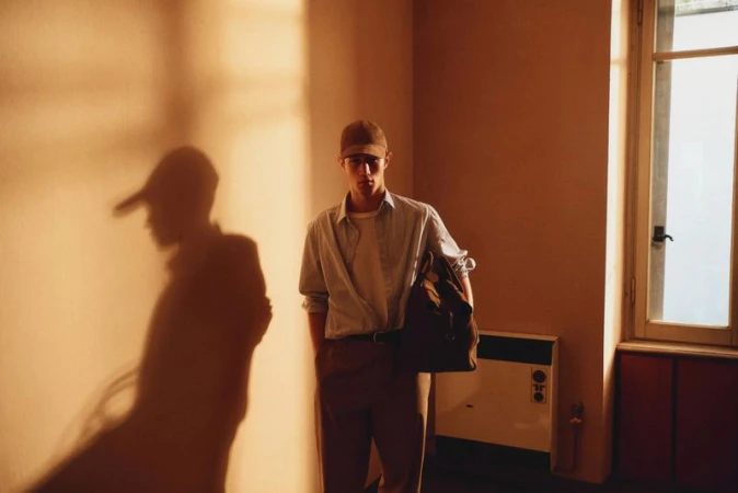 Indoor portrait of a young adult male wearing a cap, light-blue button-down shirt, and brown pants, carrying a bag, with soft lighting.