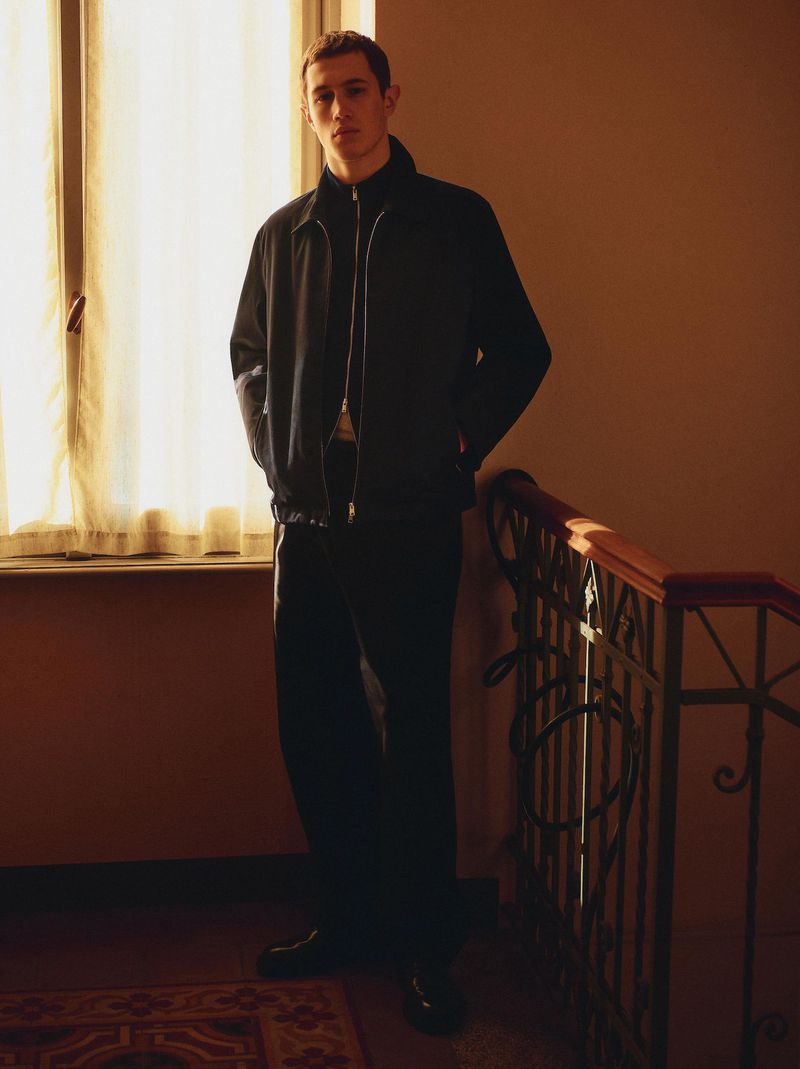 Adult male in black jacket and pants standing indoors on a staircase, near a window with natural light.