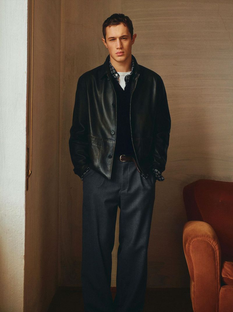 A stylish male model poses indoors wearing a black leather jacket, dark pants, and a brown belt, exuding a cool and sophisticated style.