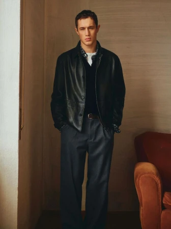 A stylish male model poses indoors wearing a black leather jacket, dark pants, and a brown belt, exuding a cool and sophisticated style.