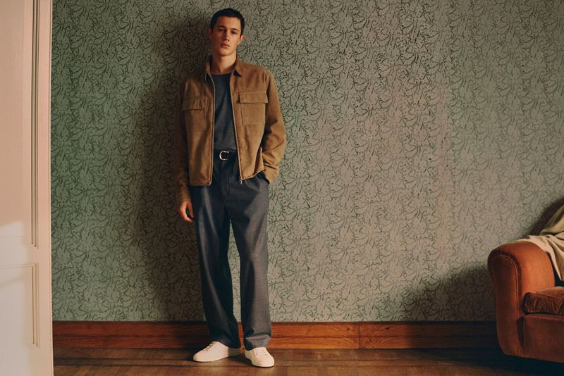 Young man in a casual brown jacket and gray pants stands against a patterned green wall in a well-lit indoor setting, posing for a fashion shot.