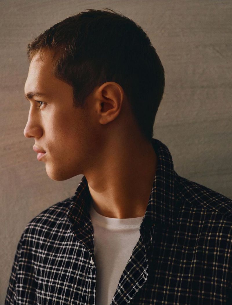 Profile shot of a fair-skinned young adult male with short brown hair wearing a black and white plaid shirt and white t-shirt, looking left.