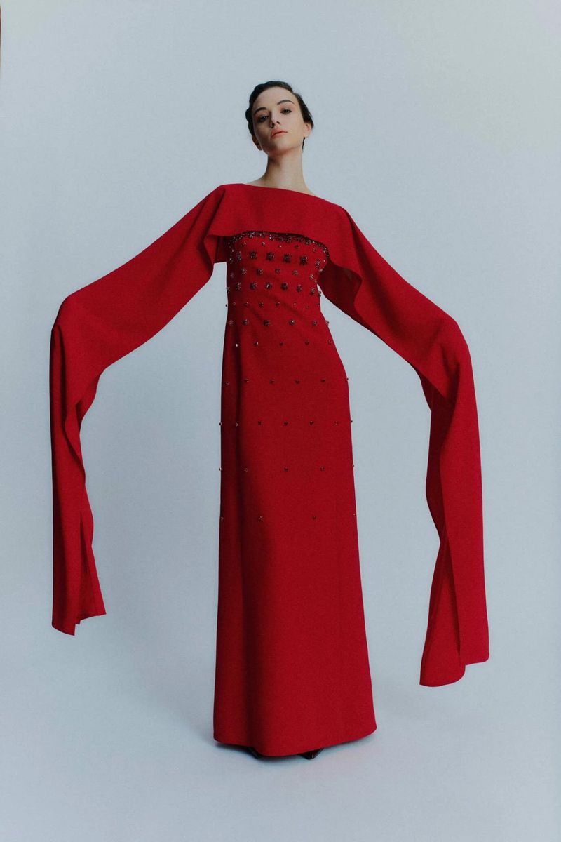 A slender woman poses in a floor-length red gown with long, flowing sleeves against a white background, showcasing a stylish fashion statement.