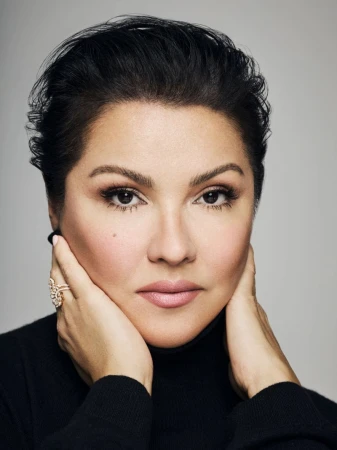 Close-up portrait of a woman with short dark hair, wearing a black turtleneck, with hands near her face, against a grey backdrop.