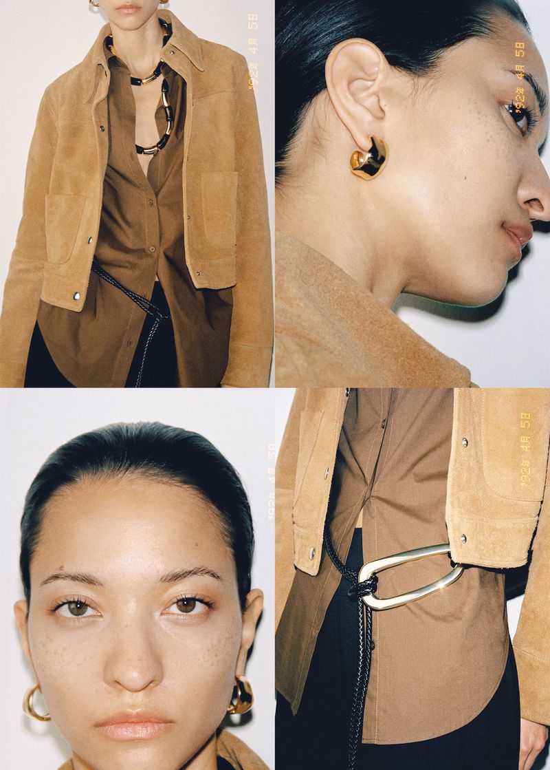 Fashion editorial shot of a woman with gold hoop earrings, wearing a brown shirt and jacket. Dark hair and a slim build are apparent.