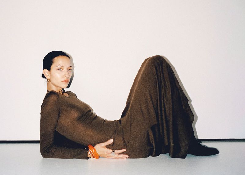 Adult woman in long-sleeve brown dress and bracelets poses lying down against white backdrop in studio lighting.