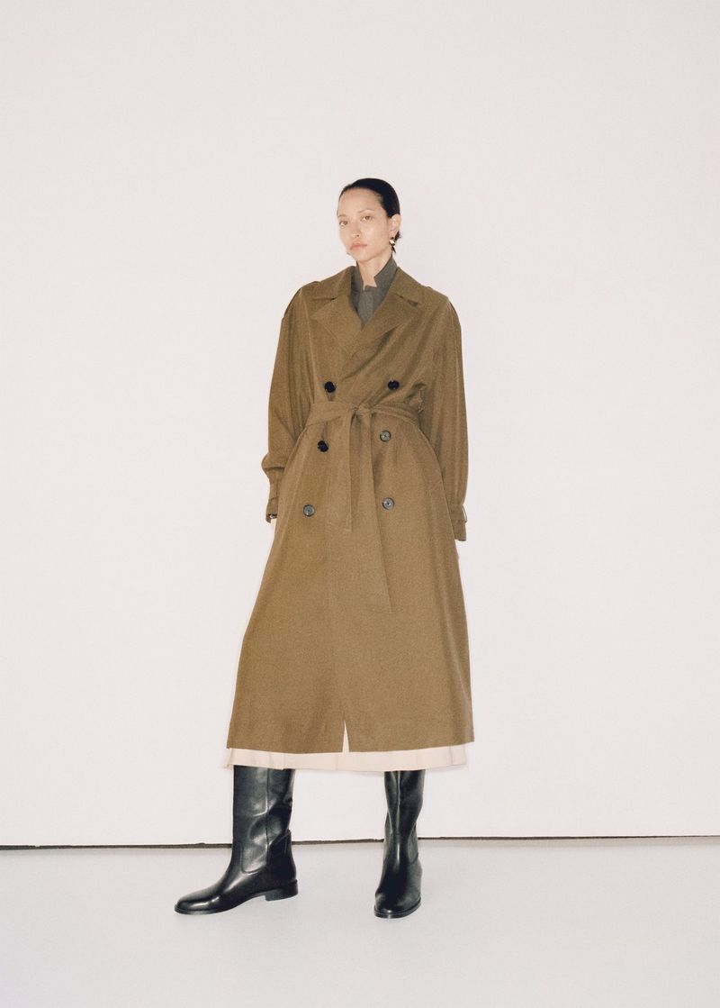 A woman wearing a trench coat and boots stands in a neutral photo studio. The clothing and model are centered in the full shot.