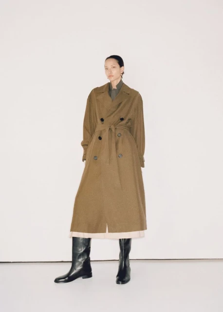 A woman wearing a trench coat and boots stands in a neutral photo studio. The clothing and model are centered in the full shot.