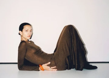 Model América González lying on a white studio floor wearing a brown long-sleeve dress and orange bangles for Mango América