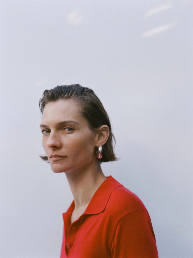 Portrait of a woman with short dark hair in a red collared shirt and silver earrings posing against a bright white background.
