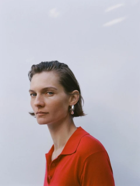 Portrait of a woman with short dark hair in a red collared shirt and silver earrings posing against a bright white background.