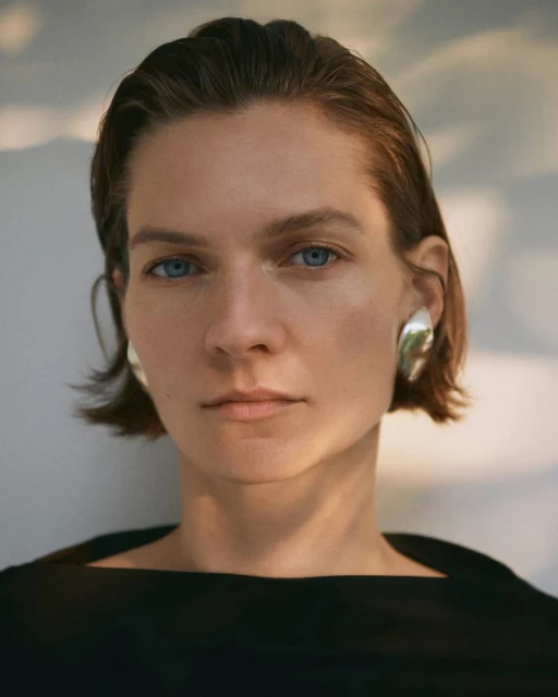 Close-up of a woman with short, slicked-back auburn hair, silver earrings, and a black dress, gazing directly at the camera.
