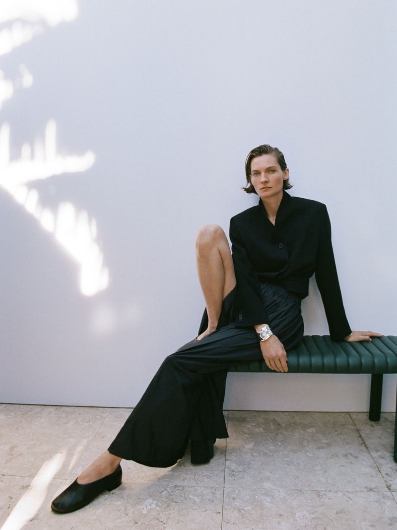 A woman in a dark blazer and wide-legged pants sits on a bench against a neutral wall with shadows, creating a fashion editorial shot.