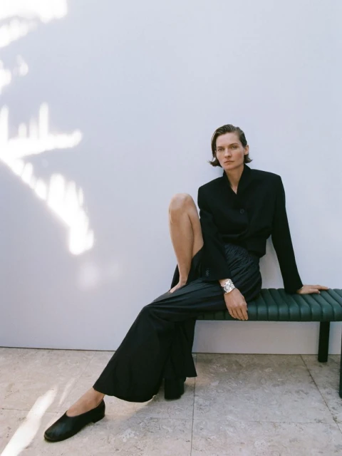 A woman in a dark blazer and wide-legged pants sits on a bench against a neutral wall with shadows, creating a fashion editorial shot.