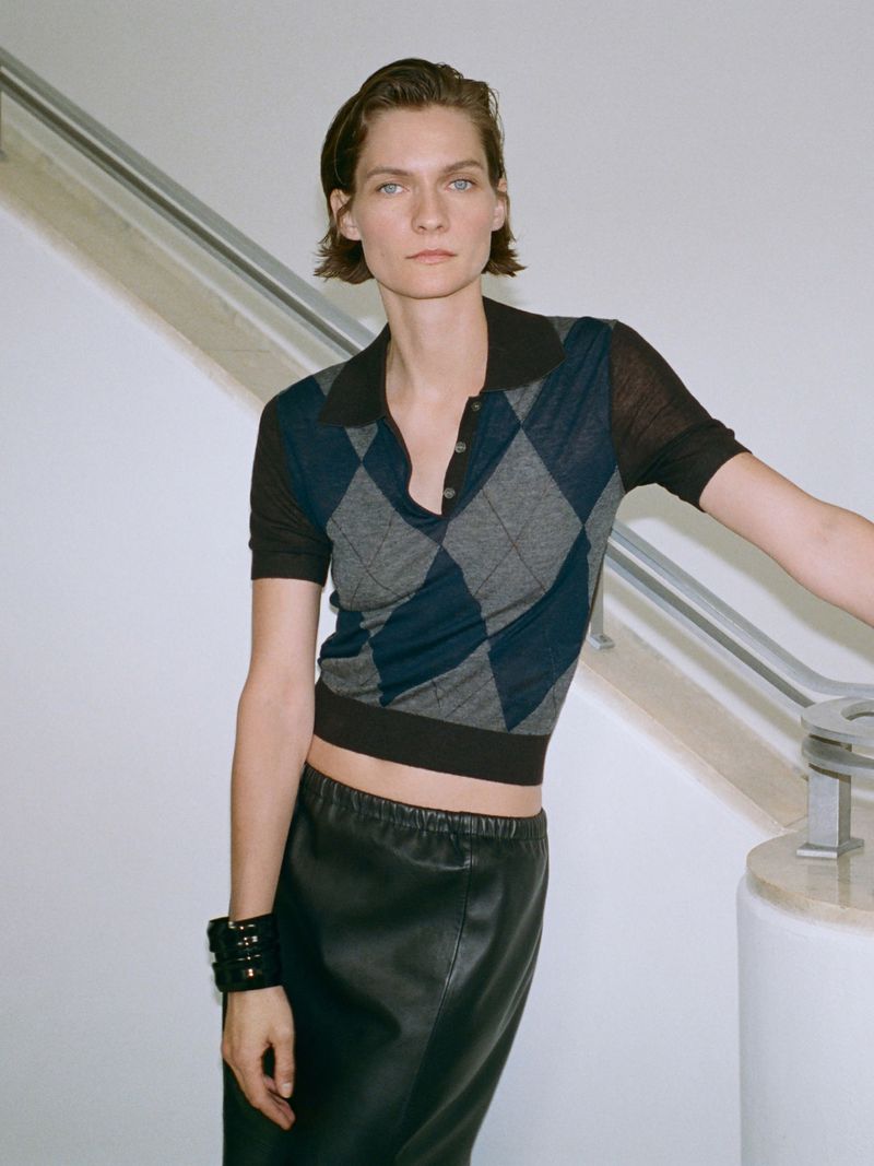 Woman in argyle polo shirt and black skirt poses indoors near white wall and stairs, looking directly at the camera with a neutral expression.