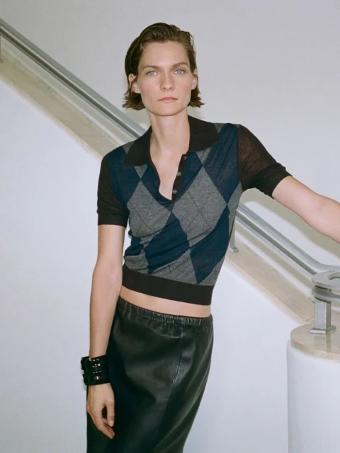 Woman in argyle polo shirt and black skirt poses indoors near white wall and stairs, looking directly at the camera with a neutral expression.