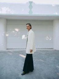Model wearing a white coat and dark trousers standing outdoors against a pale building exterior with dappled sunlight.