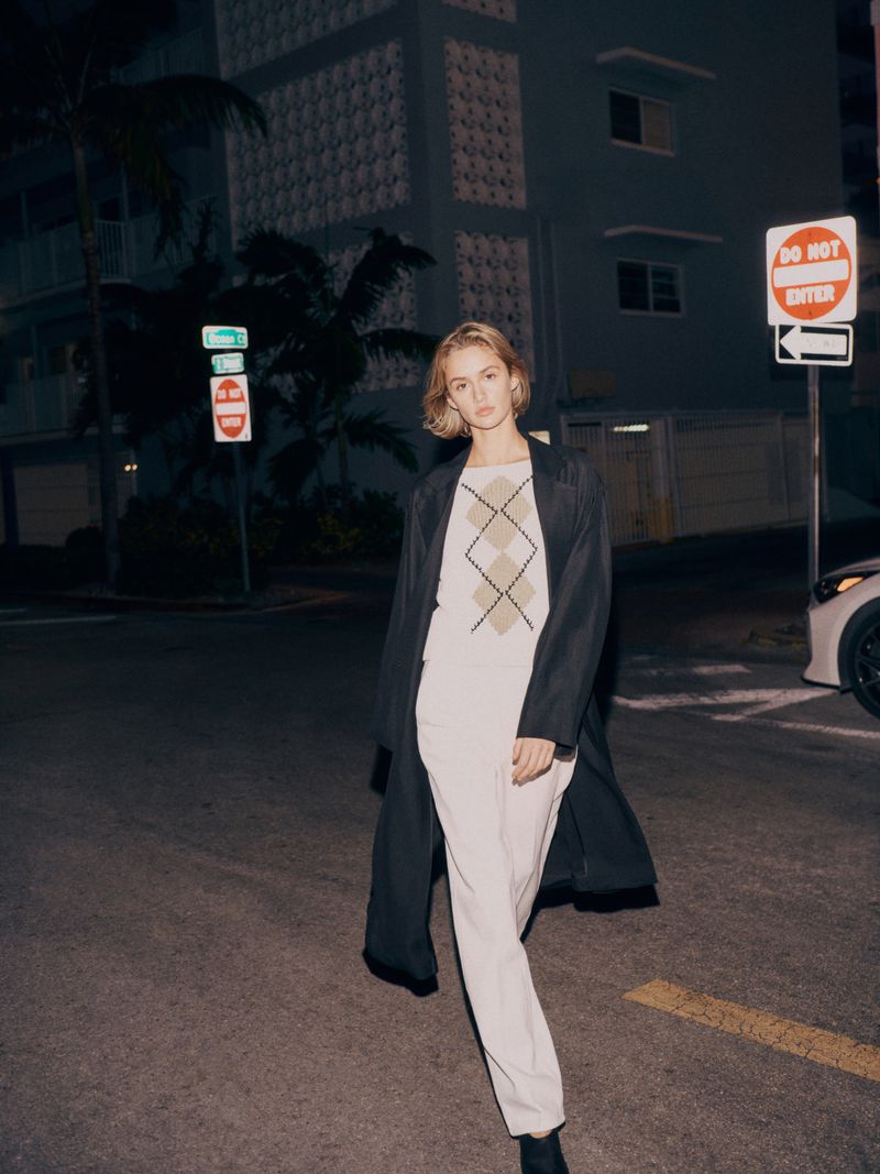 Woman walking on a city street wearing a coat and cream-colored suit with geometric-patterned top under street lights at night.