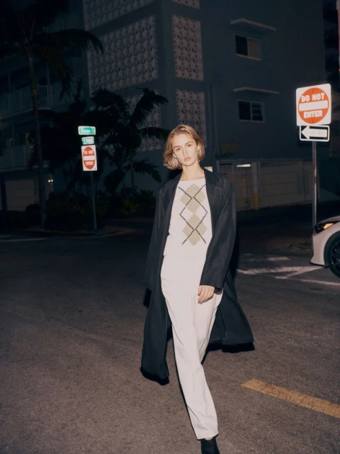 Woman walking on a city street wearing a coat and cream-colored suit with geometric-patterned top under street lights at night.