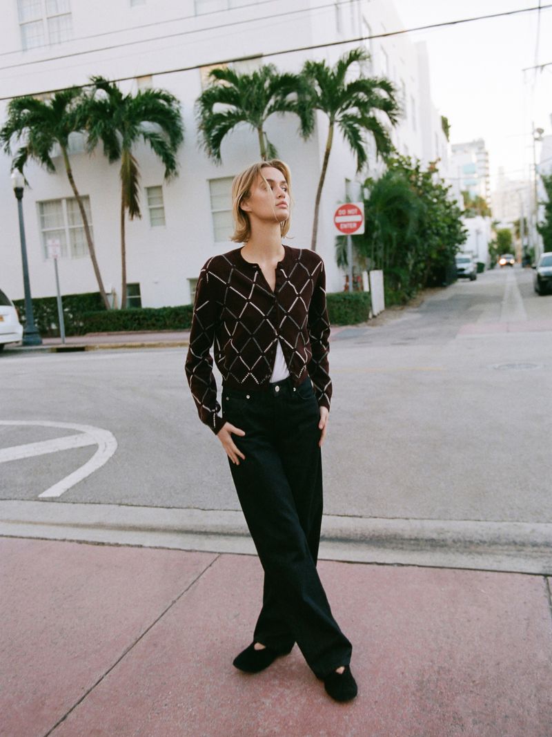 A young adult woman poses on a city street wearing a cardigan, jeans, and flats against a backdrop of white buildings and palm trees.