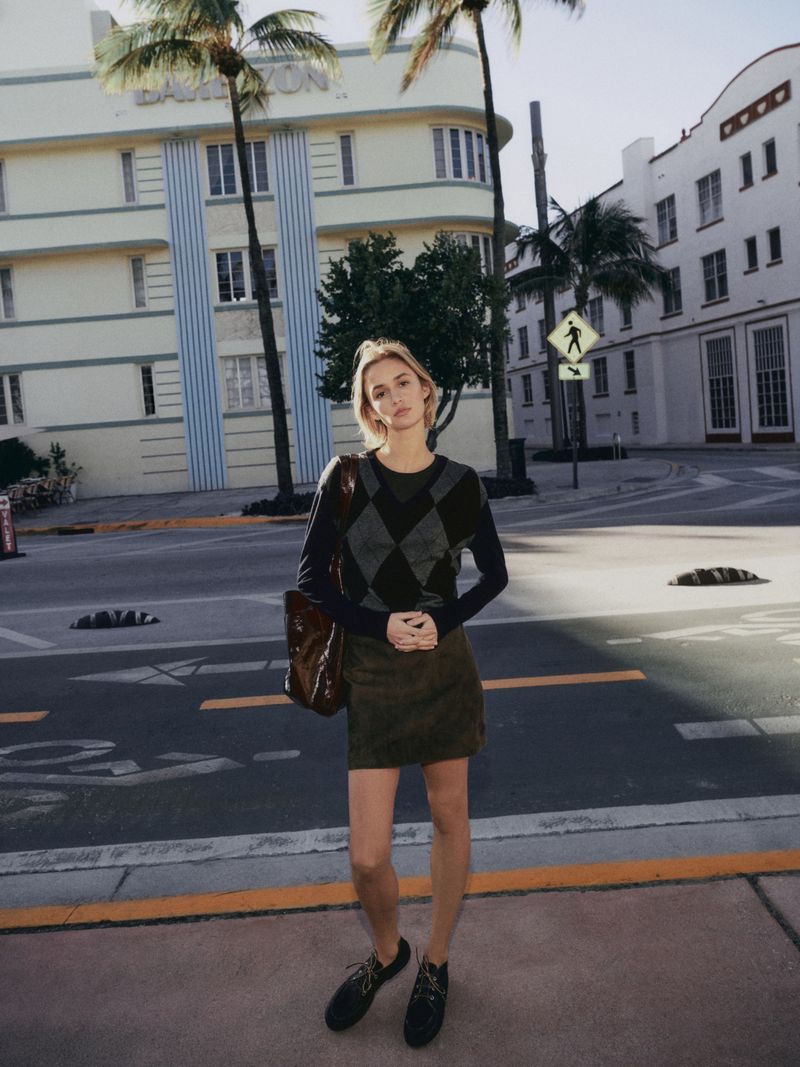 A woman stands on a sunny street in an argyle sweater and miniskirt, holding a tote bag, with palm trees and buildings in the background.