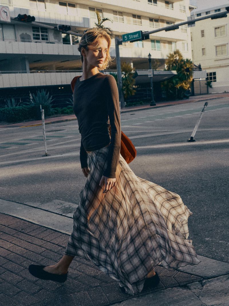 Young woman with blonde hair wearing a long-sleeve top and plaid skirt walks across a crosswalk in an urban environment on a sunny day.