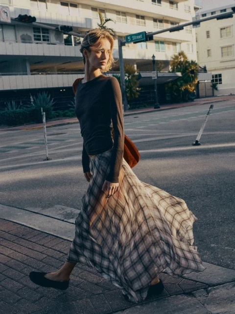 Young woman with blonde hair wearing a long-sleeve top and plaid skirt walks across a crosswalk in an urban environment on a sunny day.