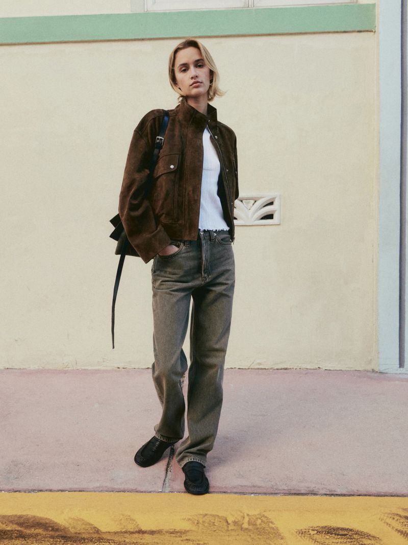 A woman stands in front of a wall wearing a brown jacket, white top, grey denim jeans, and black shoes with a black bag.