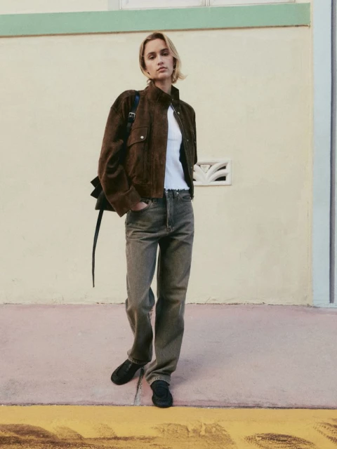 A woman stands in front of a wall wearing a brown jacket, white top, grey denim jeans, and black shoes with a black bag.