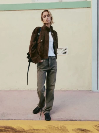 A woman stands in front of a wall wearing a brown jacket, white top, grey denim jeans, and black shoes with a black bag.