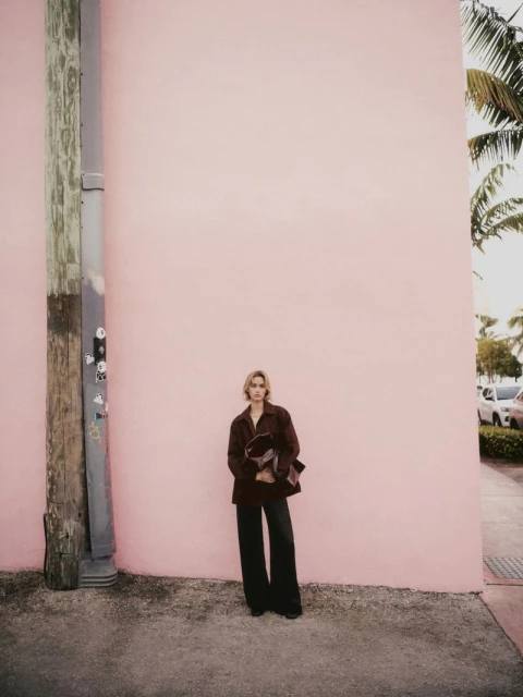 A slender adult poses against a pink wall outdoors in a dark brown jacket and dark pants, standing next to a wooden utility pole.