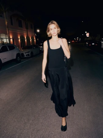 Full-length shot of a blonde woman in a black tank top and long black skirt walking on a city street at night with cars and buildings.