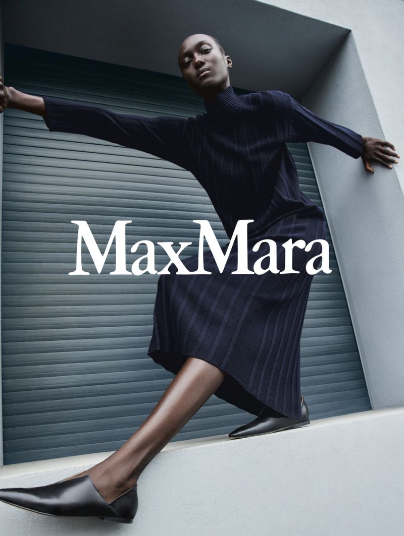 Fashion shoot of an African female adult model wearing a navy dress and black flats, leaning against a gray wall with 'MaxMara' text overlay.
