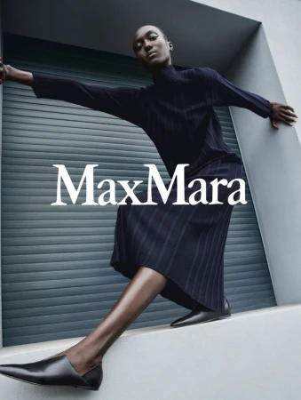 Fashion shoot of an African female adult model wearing a navy dress and black flats, leaning against a gray wall with 'MaxMara' text overlay.