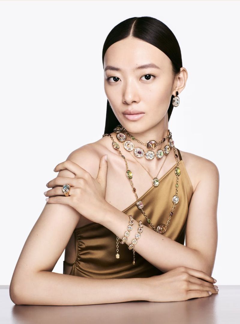 Asian woman poses in gold dress wearing ornate gold, jewel-toned necklace, bracelet, ring and earrings against a white backdrop.