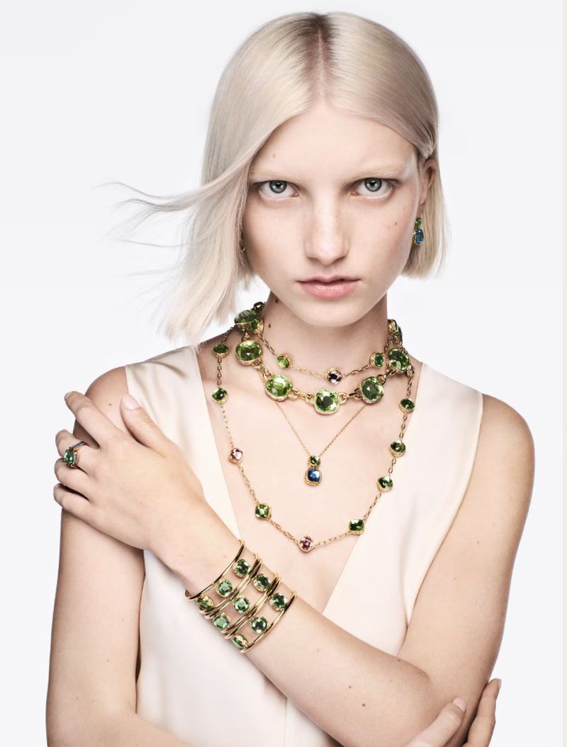 Blonde woman poses with green and gold jewelry, including a bracelet, ring, earrings, and layered necklaces against a white background.