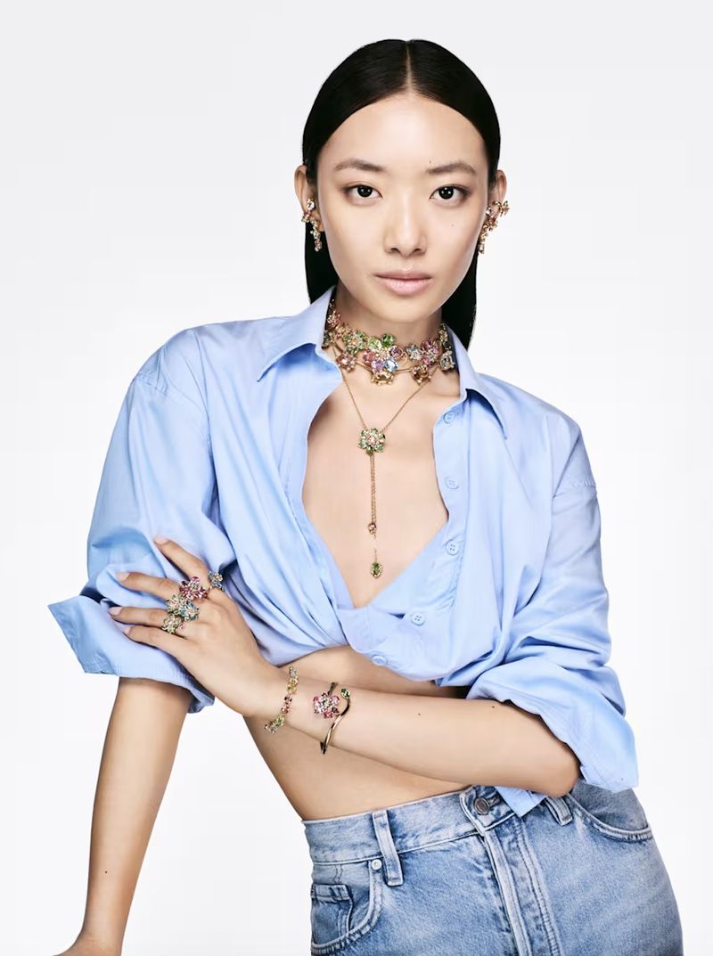 Elegant Asian woman posing indoors wearing a light blue shirt, jeans, and colorful jeweled jewelry in a beauty and fashion editorial.
