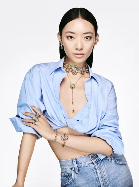 Elegant Asian woman posing indoors wearing a light blue shirt, jeans, and colorful jeweled jewelry in a beauty and fashion editorial.