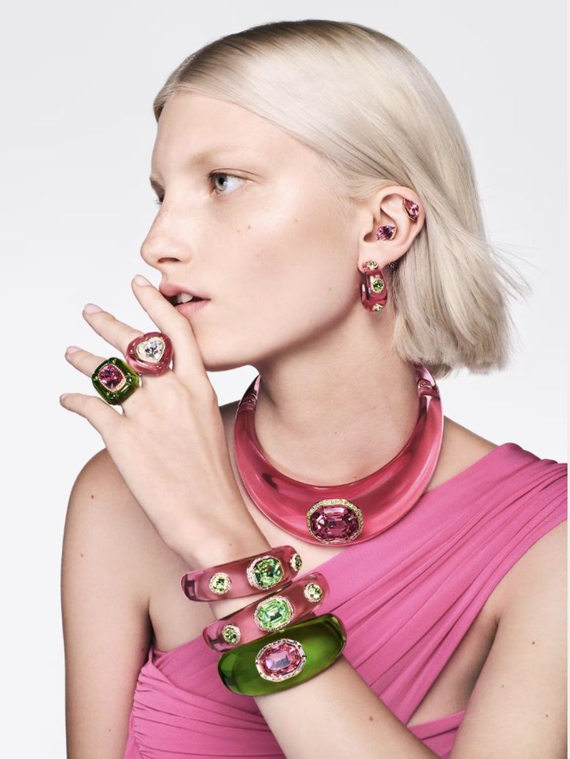 Close-up of a woman with platinum blonde hair wearing pink and green jewelry, including a necklace, bangles, rings, and earrings.