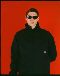 Model Valentin Humbroich wearing dark sunglasses and a black zip-up jacket against a vibrant red backdrop.