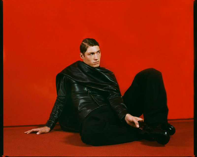 Man in black clothing poses in a studio with a red background. The model is reclined on the floor with one hand resting on the ground.