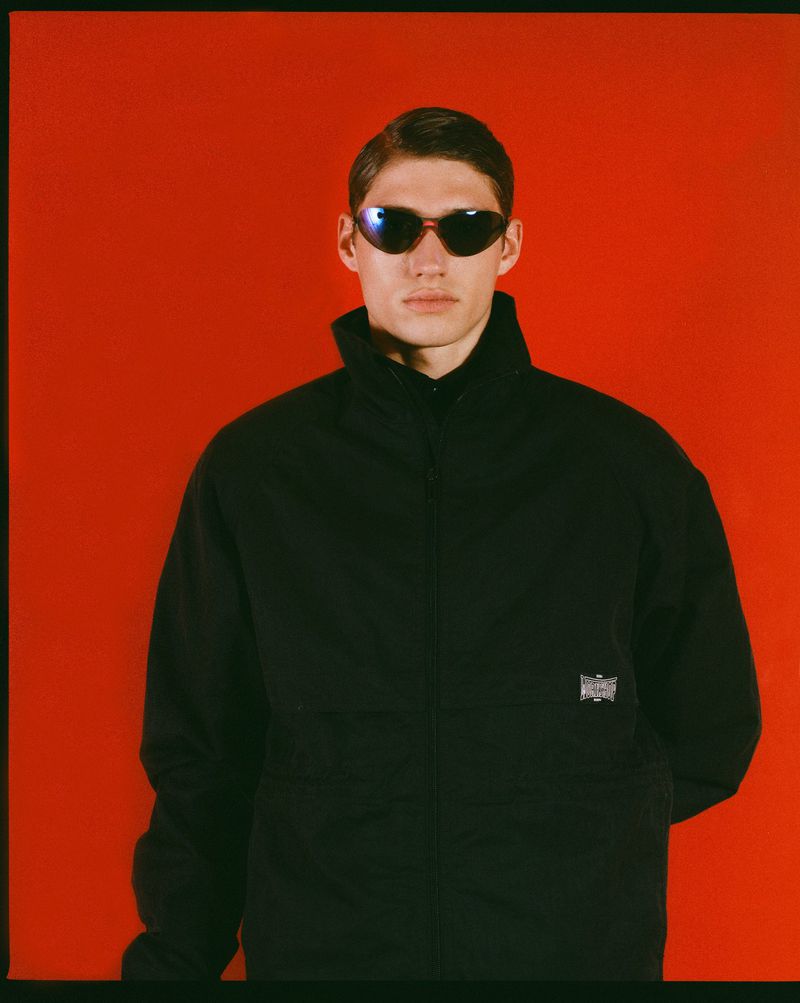 Portrait of a young adult man in a black jacket and sunglasses posing against a vibrant red background in a fashion-style photograph.